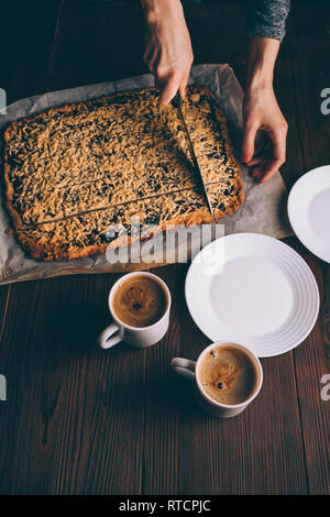 Die Hände oben Frau schneiden frisch gebackene Plätzchen neben zwei Tassen Kaffee und Platten auf braune Holztisch. Stockfoto