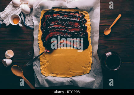 Teig mit gespreizten Johannisbeere Konfitüre auf Backpapier Neben Eiern, Muscheln und Eigelb gerollt. Ansicht von oben Prozess der Herstellung hausgemachte Kuchen auf braunem Holz- t Stockfoto