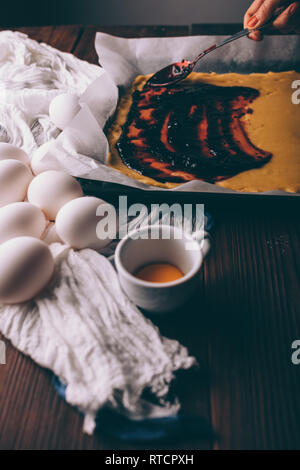 Prozess der Herstellung hausgemachte Kuchen. Nahaufnahme der Hand des weiblichen Verbreitung Johannisbeere Stau auf gewalzten Teig neben Eier und Eigelb auf braune Holztisch. Stockfoto