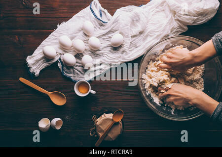 Blick von oben Die weibliche Hände kneten den Teig in der grossen Schüssel in der Nähe von Zutaten und Utensilien für die Küche hausgemachte Kuchen auf braun Küche aus Holz Tisch. Stockfoto