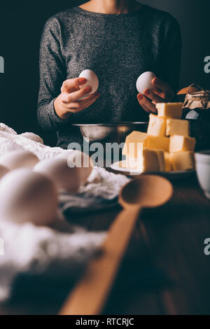 Frau sitzt am Tisch, die Eier in der Nähe von Zutaten und Utensilien zum Kochen Kuchen: gewürfelte Butter, Marmeladenglas und Löffel aus Holz. Stockfoto