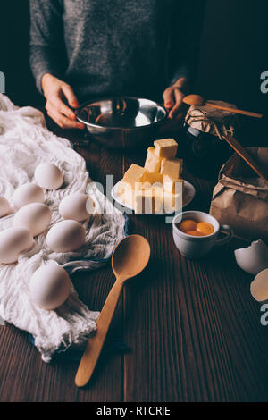 Prozess der Herstellung hausgemachte Kuchen auf braune Holztisch. Zugeschnittenes Bild Frau mit Metall Schüssel in der Nähe von Backen Zutaten: Eier, Eigelb, gewürfelte Butter und Fl Stockfoto