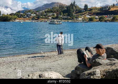 Asiatische und westliche Touristen und Einheimischen stehen und lesen Buch auf dem Kiesstrand an Sommertag im Sonnenschein Lake Wanaka Neuseeland Südinsel Stockfoto