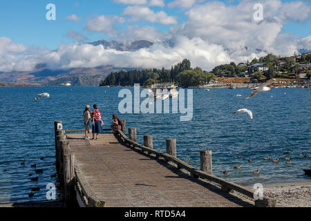 Asiatische und westliche Touristen und einheimische Vögel füttern am Kieselstrand und Pier auf der Sommertag im Sonnenschein Lake Wanaka Neuseeland Südinsel Stockfoto