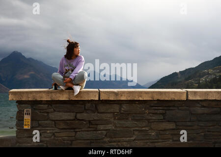 Asiatische Touristen, die eine Tasche sitzt am Strand Wand in Queenstown mit Blick auf Lake Wakatipu Neuseeland Südinsel Stockfoto