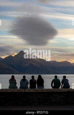 Touristen sitzen am Strand Wand den Sonnenuntergang über den Lake Wakatipu, Queenstown, Neuseeland Südinsel Stockfoto