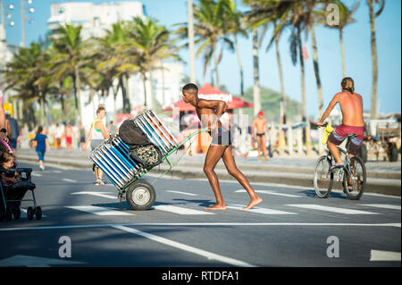 RIO DE JANEIRO - 26. MÄRZ 2017: Eine junge brasilianische Arbeiter schiebt einen Einkaufswagen der gefalteten Stühle zum Strand stehen auf der Straße am Strand in Ipanema. Stockfoto