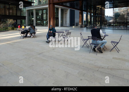 Menschen entspannend und mit Hilfe eines Handys an den Tischen in einem Hof in der Nähe von Kings Cross, London, England sitzen Stockfoto