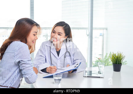 Ärztin spricht mit weiblichen Patienten im Krankenhaus Büro beim Schreiben auf die Patienten Gesundheit Datensatz in der Tabelle. Gesundheitswesen und medizinische Betreuung. Stockfoto