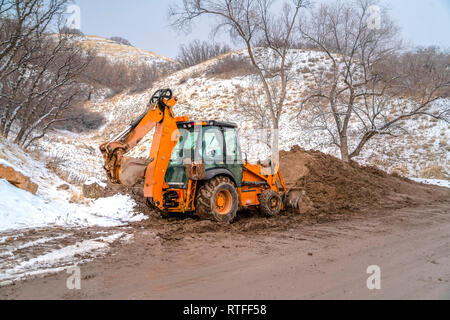 Baggerlader gegen verschneite Hügel und Bäume. Eine orange Baggerlader auf der Seite einer Straße mit einem erdhügel Boden geparkt. Hügel und Bäume mit s abgedeckt Stockfoto