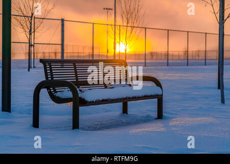 Verschneite Bank mit Sonnenuntergang im Hintergrund. Eine verschneite Bank auf schneebedeckten Boden in Eagle Mountain, Utah bei Sonnenuntergang. Die untergehende Sonne leuchtet in Stockfoto