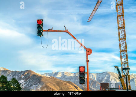 Ampel und Kran gegen Berg und Himmel. Viw Ampel und ein gelber Bau tower Kran an einem sonnigen Tag. Verschneite Berge und Clou Stockfoto