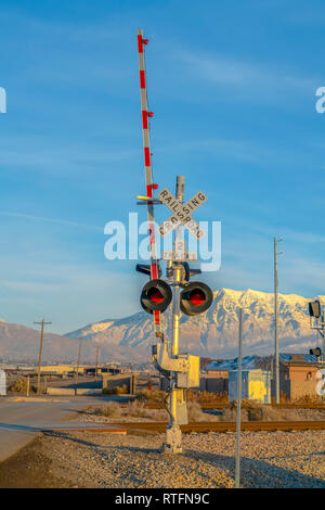 Zwei tracks Bahnübergang neben einer Eisenbahn. Zwei tracks Bahnübergang mit Schranke und roten Ampeln. Gebäude, Berg und Himmel werden kann Stockfoto