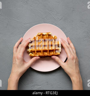 Ansicht von oben, weibliche Hände, die traditionelle belgische Waffel auf Rosa die Platte über der Betonoberfläche. Overhead, von oben. Stockfoto