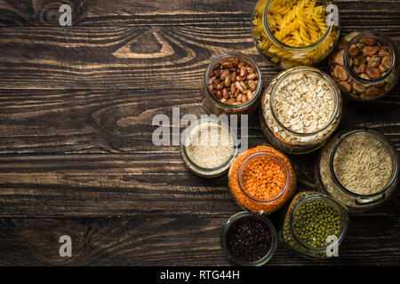 Getreide, Hülsenfrüchte und Bohnen in Gläsern auf dunklen Holztisch. Stockfoto