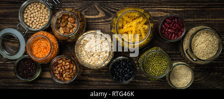 Getreide, Hülsenfrüchte und Bohnen in Gläsern auf dunklen Holztisch. Stockfoto