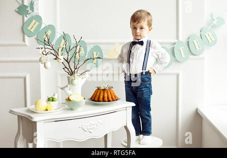 Süße Junge in ein weißes Hemd und Quaste Schmetterling steht auf einem weißen Stuhl neben einer Tabelle in einem weißen Raum auf einem Tisch. Ostern Dekoration einer Vase mit branc Stockfoto