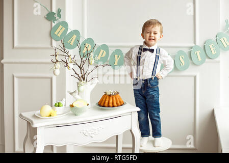 Süße Junge in ein weißes Hemd und Quaste Schmetterling steht auf einem weißen Stuhl neben einer Tabelle in einem weißen Raum auf einem Tisch. Ostern Dekoration einer Vase mit branc Stockfoto