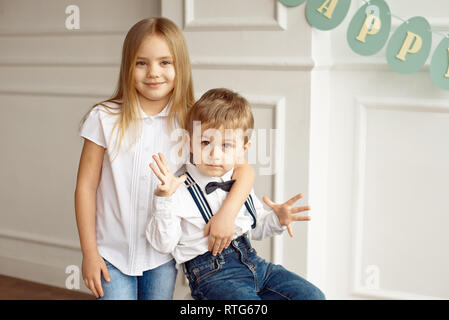 Süße Junge in einem weißen Hemd sitzt auf einem Stuhl in einem hellen Zimmer Schwester umarmt seine Schultern Stockfoto