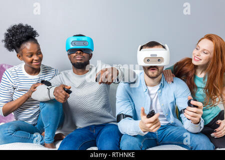 Zwei Männer unterschiedlicher ethnischer Herkunft tragen VR-Headset, spielen in Game Studio und ihre Freundinnen sie unterstützt, neben sitzen auf der Couch und Chee Stockfoto