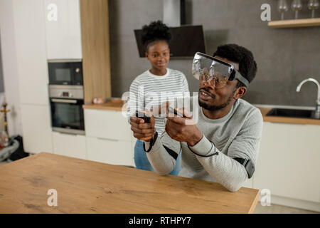 Afrikanischer mann Vornehmen der Einstellungen der virtuellen Realität Konsole mit Controller, während seine Frau ihm die Vorbereitung am Morgen Kaffee in der modernen Küche Stockfoto