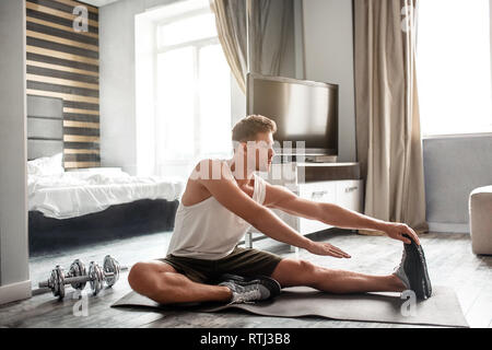 Die Jungen gut gebauten Mann gehen in für den Sport in der Wohnung. Er sitzen auf carimate und elastischer Körper. Guy erreichen Ende der Fuß mit der Hand. Muskulöse Mann konzentriert Stockfoto