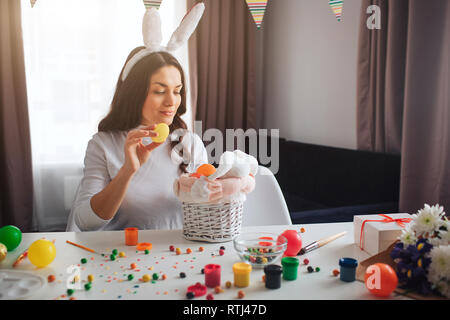 Junge hübsche Frau, die Vorbereitung auf Ostern. Sie sitzen und dekorieren Warenkorb und Kaninchen Spielzeug mit bunten Eiern. Dekoration und Süßigkeiten auf dem Tisch. Modell tragen Rabbi Stockfoto