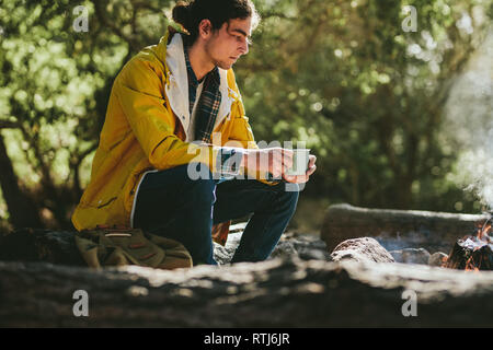 Seitenansicht eines Mann neben einem Lagerfeuer Holding eine Kaffeetasse sitzend. Junger Mann tief in Gedanken am Lagerfeuer. Stockfoto