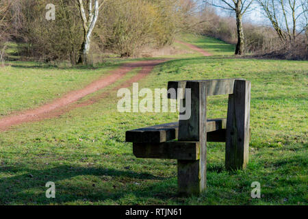 Eine hölzerne Parkbank im grünen Gras sitzt neben einem Feldweg umgeben von Bäumen Stockfoto