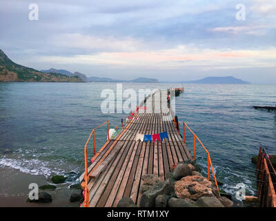 Perspective view of a wooden pier mole at sea. Wooden bridge in spring time with blue sky. Place for fishing with pier. Dark and Foggy sea with hills Stockfoto