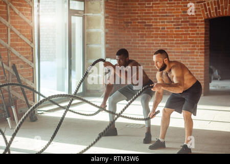 Zwei multiethnischen atletic Freunde der Stärke von einem unsichtbaren Feind mit Schlacht Seil in crossfit style Fitness Test, stehend mit Beine auseinander, Schulter - wid Stockfoto