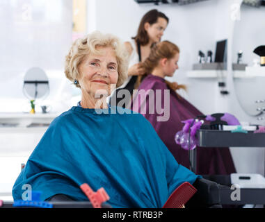 Elegante ältere Frau in Friseursalons Cape in Friseur Stuhl Sitzen, Warten auf Restaurantbesuche Stockfoto