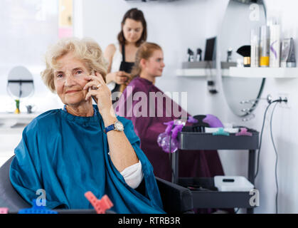 Elegante ältere Frau in Friseursalons Kap sitzen in Friseur Stuhl mit Telefon während der Wartezeit für Restaurantbesuche Stockfoto