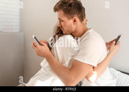 Junges Paar im Schlafzimmer nach dem Aufwachen. Seitenansicht des untreuen Mann und Frau sind Betrug am Telefon, während Umarmen Stockfoto