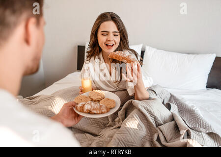 Junges Paar mit einem köstlichen Frühstück im Bett nach dem Aufwachen. Schöne Frau ist Essen frisches Croissant mit Saft Stockfoto