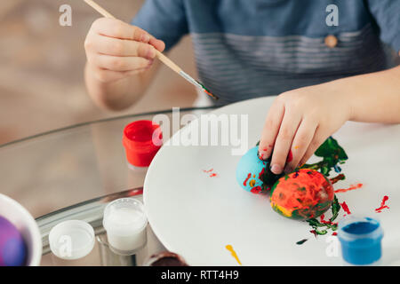 Hausgemachte Ostern Dekorationen und kleinen Helfer. Selektiver Fokus der kleinen Hände eines Jungen, gekochte Eier mit bunten Farben auf weißem Schild Stockfoto