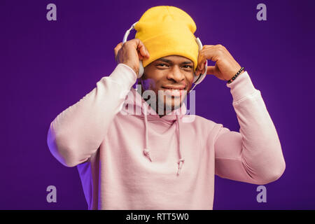 Junge afrikanische Mann in Hoodie, in der Sinn für Mode eines street Urchin, drehte sich ein wenig in der Kopfhörer auf der violette Hintergrund isoliert, holding Ohrhörer Stockfoto