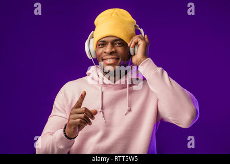 Junge afrikanische Mann in Hoodie, in der Sinn für Mode eines street Urchin, drehte sich ein wenig in der Kopfhörer auf der violette Hintergrund isoliert, holding Ohrhörer Stockfoto