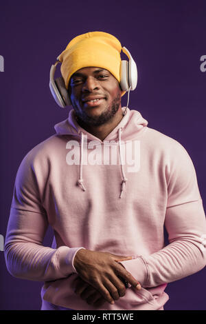 Junge afrikanische Mann in Hoodie, in der Sinn für Mode eines street Urchin, drehte sich ein wenig in der Kopfhörer auf der violette Hintergrund isoliert, holding Ohrhörer Stockfoto