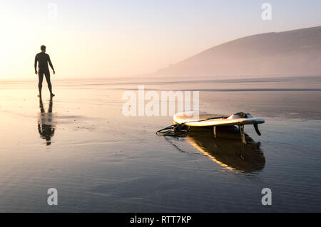 Ein einsamer Surfer mit Blick auf das Meer. Mit dem Surfbrett spiegelt sich auf dem nassen Sand. Saunton Sands, Devon, Großbritannien. Am wärmsten Februar auf Aufzeichnung. 27.0 Stockfoto