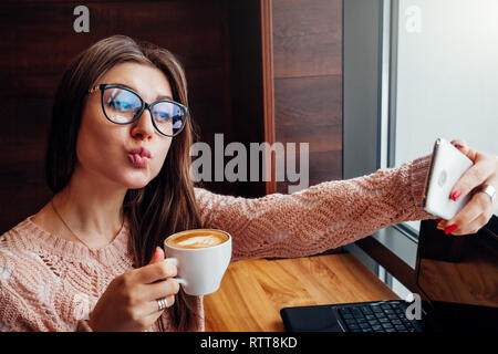 Junge hübsche Frau Freelancer macht einen selfie und genießt Kaffee im Cafe vor dem Fenster Stockfoto