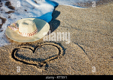 Ein Bild von einem Herz am Strand mit einem staw Hut und kommenden Welle. Platz für Valentinstag text Stockfoto