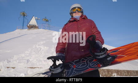 Ältere Frau mit dem Snowboard. Ansicht von unten gerne ältere Frau in Helm und Brille, tragen Skianzug, stand vor der Skilift mit einem Snowboard Stockfoto