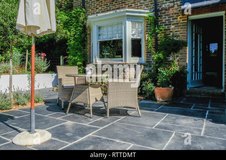 Korbstühle und Rohrgeflecht Tisch in der Mitte eines Schiefer Patio mit Pflanzen und Bäumen in Beeten und Töpfen. An einem sonnigen Sommertag mit niemand in der übernommen Stockfoto