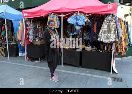 Eine lächelnde Frau verkaufen afrikanischen Textilien, Kleidung und Schmuck zu einem Straßenmarkt auf Brixton Road in Brixton South London UK KATHY DEWITT Abschaltdruck Stockfoto