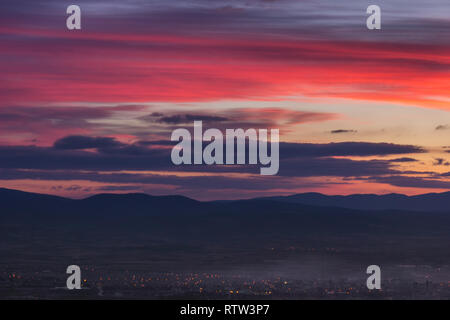 Dramatische, bunte spät Blaue Stunde Himmel über weit entfernte Stadt in Nebel und Silhouette Horizont Berge bedeckt Stockfoto