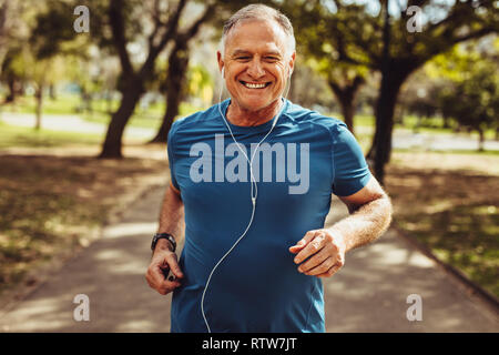 Portrait eines älteren Mannes in fitness Verschleiß in einem Park läuft. Nahaufnahme von einem lächelnden Mann laufen beim Hören von Musik über Kopfhörer. Stockfoto