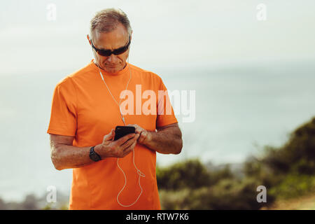 Fitness-Mann, der während des Workouts Musik auf dem Mobiltelefon spielt. Nahaufnahme eines athletischen älteren Mannes mit dunkler Brille, der sein Handy überprüft Stockfoto