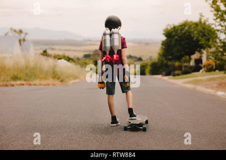 Ansicht der Rückseite des Junge mit Helm und Spielzeug Jetpack stehend mit seinem Skateboard und suchen die Straße hinunter. Jetpack Junge mit Skateboard Stockfoto