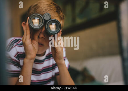 Kleiner Junge auf der Suche außerhalb Fenster mit Fernglas. Junge Blick durch ein Fernglas aus dem Wohnzimmer. Stockfoto